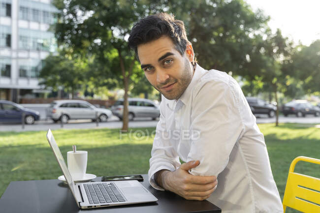 Hombre de negocios seguro con portátil en la mesa sentado en la cafetería de la acera - foto de stock