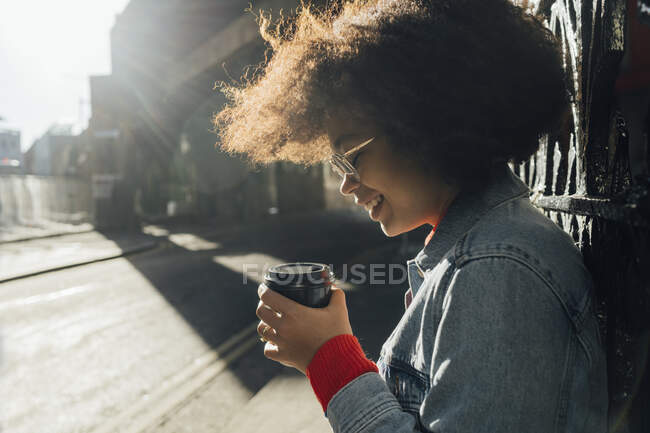 Close-up de sorrindo afro mulher segurando copo descartável na rua durante o dia ensolarado — Fotografia de Stock