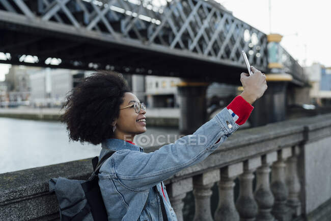 Afro giovane donna prendere selfie con smart phone mentre in piedi da ringhiera in città — Foto stock