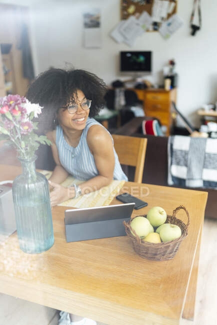 Mujer de negocios sonriente con tableta digital en el escritorio mirando hacia otro lado mientras se sienta en casa - foto de stock