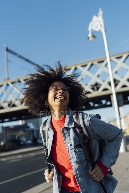 Joyeux jeune femme aux cheveux afro debout contre un pont en ville par une journée ensoleillée — Photo de stock