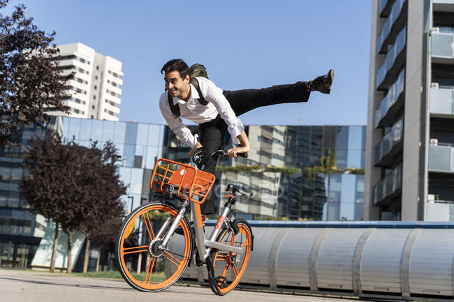 Беззаботный мужчина-предприниматель прыгает на электрическом велосипеде против ясного неба в городе — стоковое фото