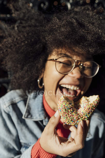 Close-up of afro young woman holding donut laughing outdoors during sunny day — Stock Photo