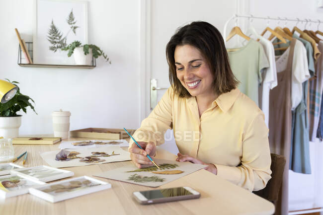 Belle femme souriante peignant avec des feuilles sur du carton tout en étant assise à table — Photo de stock
