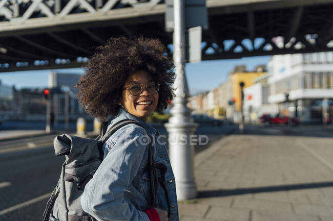 Mujer joven sonriente con cabello afro mirando hacia otro lado mientras está de pie en la acera durante el día soleado - foto de stock