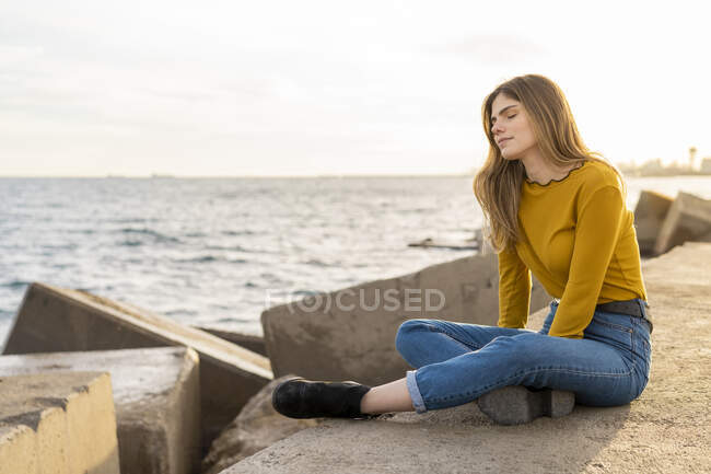 Jeune femme avec les yeux fermés assis et jouissant près de la mer contre le ciel — Photo de stock