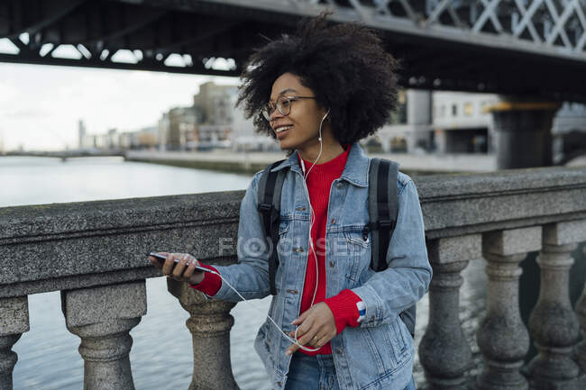 Afro giovane donna ascoltare musica attraverso le cuffie mentre in piedi da ringhiera sul ponte pedonale — Foto stock