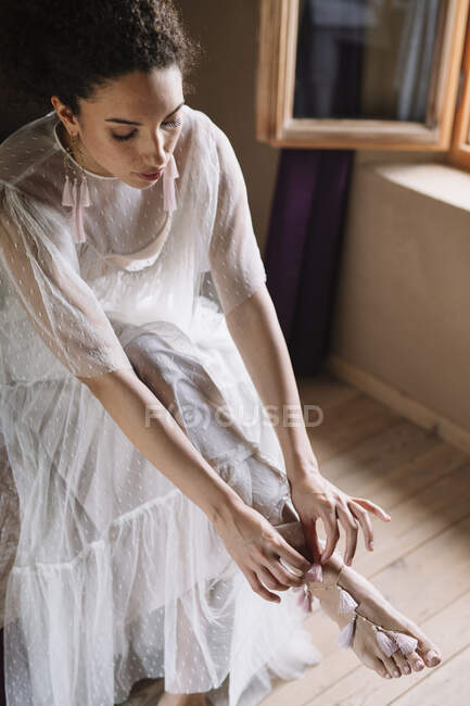 Bride wearing jewelry on leg while sitting by window at dressing room — Stock Photo
