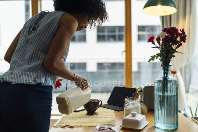 Mujer joven vertiendo leche en la taza de café en la mesa en casa - foto de stock