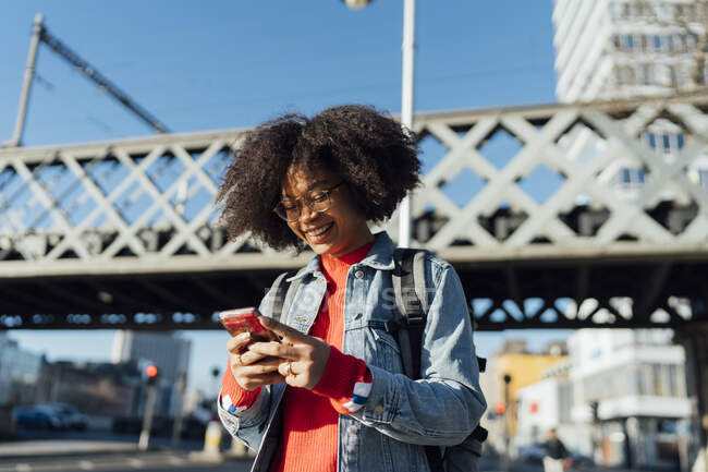 Smiling afro young woman using smart phone while standing against bridge in city — Stock Photo