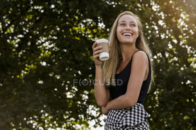 Allegro giovane donna in possesso di tazza di caffè mentre in piedi contro gli alberi nel parco — Foto stock
