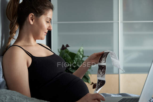 Беременная женщина проводит ультразвуковое сканирование во время использования ноутбука на кровати дома — стоковое фото
