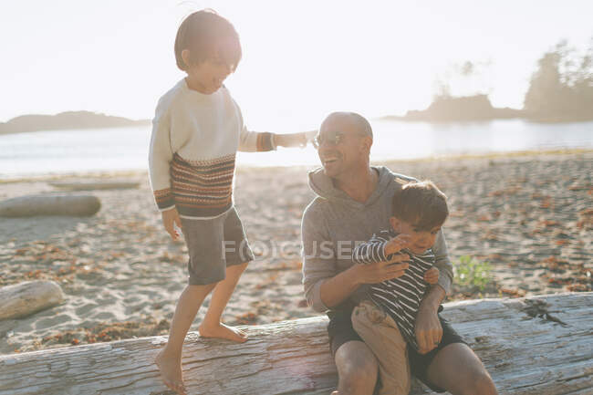 Père souriant avec des enfants assis sur du bois flotté à la plage — Photo de stock
