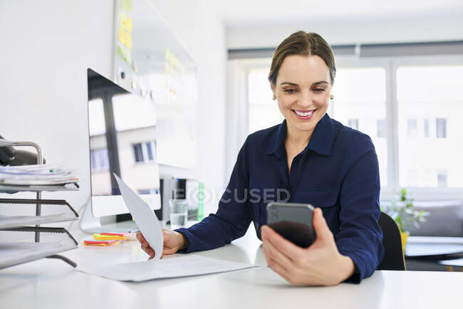 Empresária criativa usando telefone inteligente ao fazer papelada no escritório — Fotografia de Stock