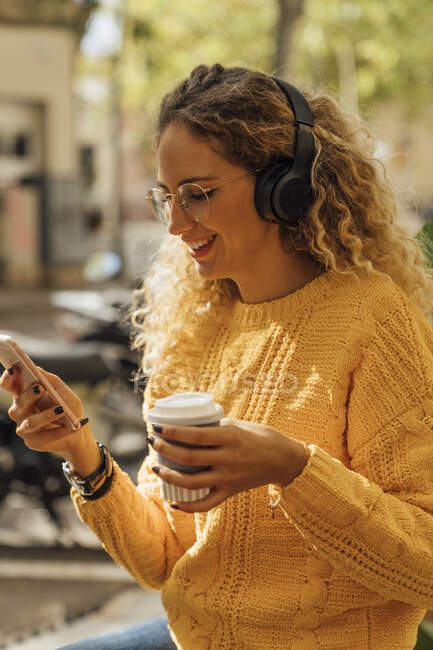 Mujer joven sonriente usando un teléfono inteligente mientras escucha música sosteniendo una taza desechable en la ciudad - foto de stock
