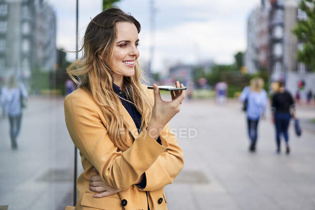 Mujer sonriente usando altavoz mientras habla a través del teléfono inteligente en la estación de tranvía - foto de stock