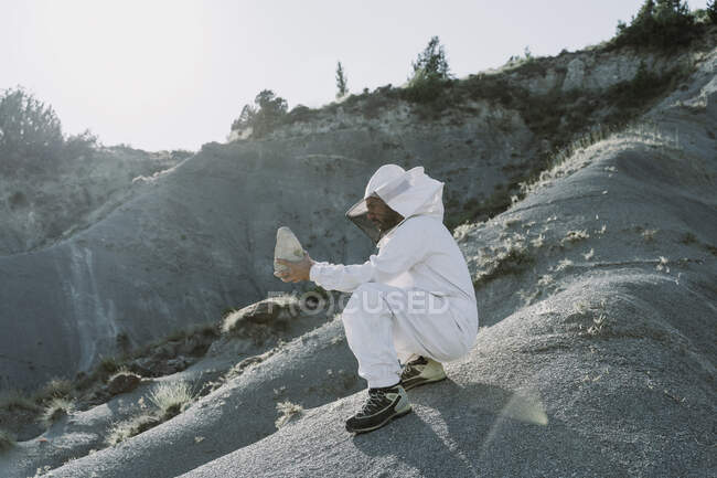 Hombre con un vestido de apicultor en un paisaje apocalíptico seco sosteniendo una piedra - foto de stock