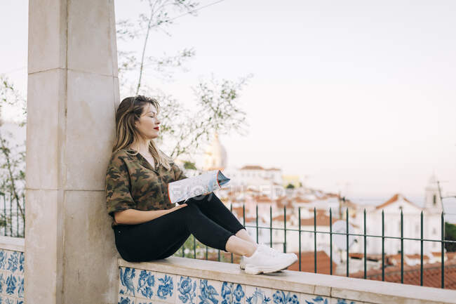 Junge Frau mit Karte sitzt auf Stützmauer gegen klaren Himmel — Stockfoto