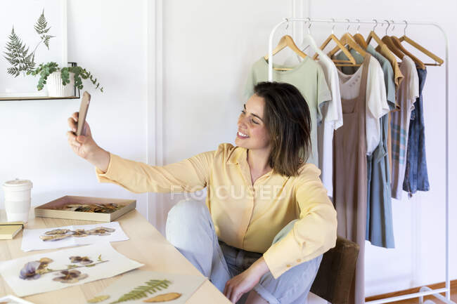 Souriant belle jeune femme prenant selfie de téléphone intelligent tout en étant assis à table avec des fleurs et des feuilles sèches — Photo de stock