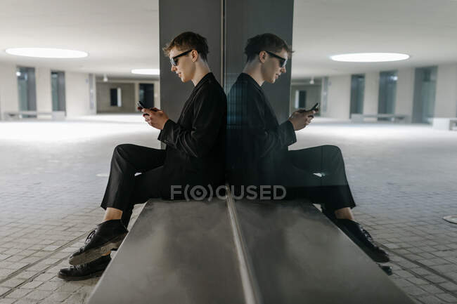 Hombre joven trans con gafas de sol negras usando teléfono móvil mientras está sentado en el asiento en el sótano - foto de stock