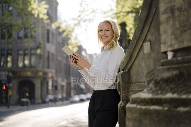 Femme entrepreneure souriante tenant une tablette numérique tout en s'appuyant sur le mur environnant et regardant loin dans la ville — Photo de stock
