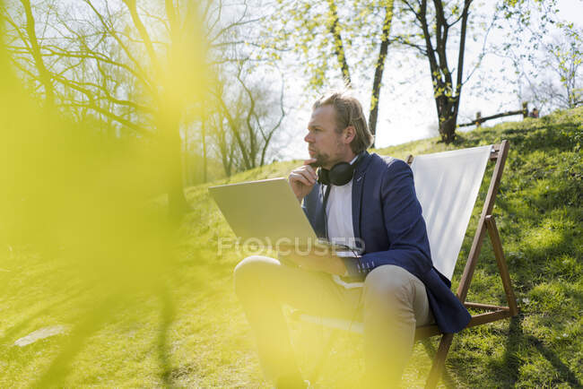 Задумчивый бизнесмен смотрит в сторону, сидя с ноутбуком в парке в солнечный день — стоковое фото