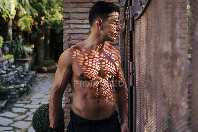 Atleta maschio senza maglietta che guarda attraverso la recinzione mentre in piedi in cortile — Foto stock