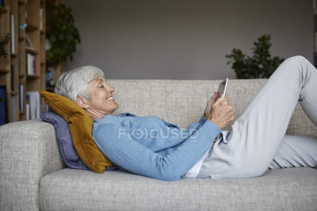 Donna sorridente che utilizza tablet digitale mentre sdraiato sul divano a casa — Foto stock