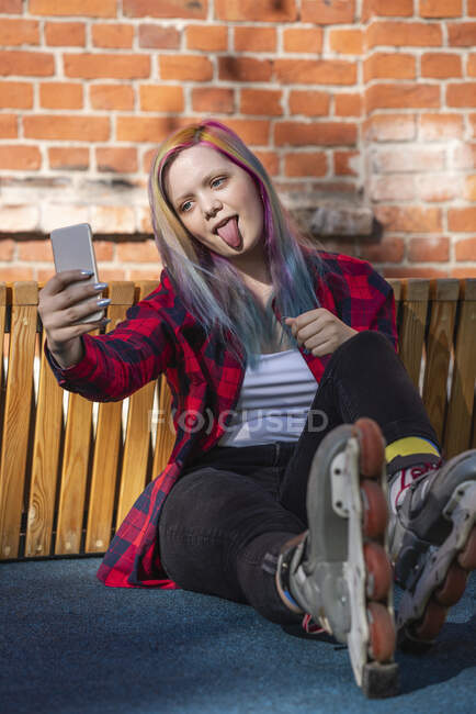 Young woman with dyed hair and roller skates taking a selfie and sticking out her tongue — Photo de stock