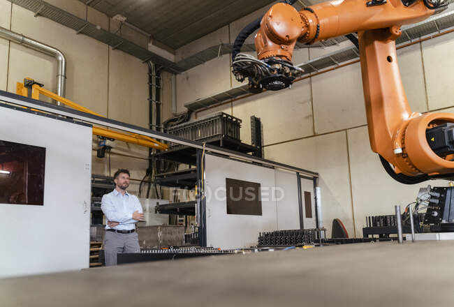 Бизнесмен со скрещенными руками контролирует автоматическую руку робота, стоя на заводе — стоковое фото