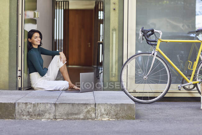 Mujer emprendedora sonriente con portátil sentada en la entrada de la oficina - foto de stock