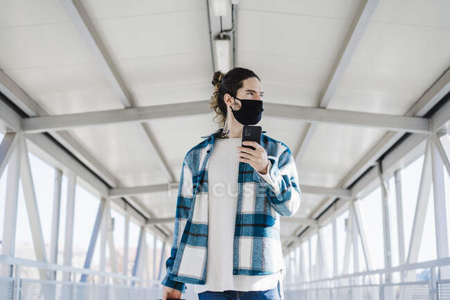 Junger Mann mit Gesichtsmaske schaut weg, während Handy auf Brücke steht — Stockfoto