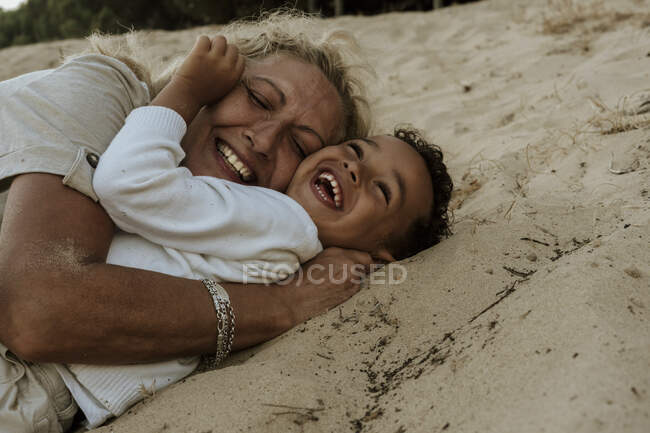 Grandmother and grandson embracing each other while lying down on sand at beach during sunset — Stock Photo
