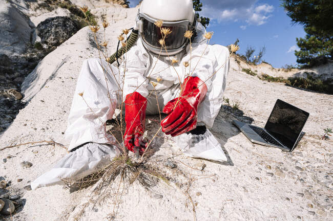 Male astronaut picking plant while sitting with laptop on mountain — Stock Photo