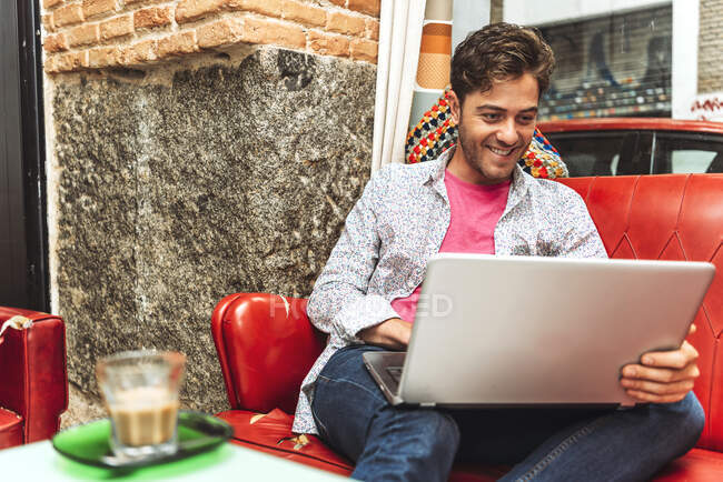 Sonriendo hombre adulto medio usando el ordenador portátil mientras está sentado en el sofá en la cafetería - foto de stock