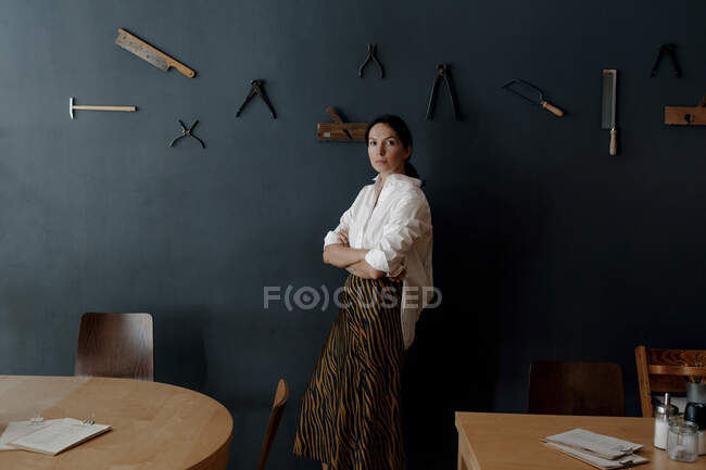 Proprietario donna con le braccia incrociate in piedi da strumento di lavoro appeso a parete in caffetteria — Foto stock
