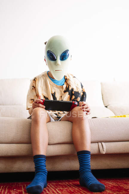 Niño usando máscara alienígena jugando videojuego de mano mientras está sentado en el sofá en casa - foto de stock