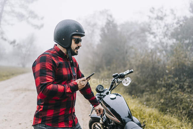 Motociclista maschio che parla in vivavoce di smartphone mentre è in piedi in moto in tempo nebbioso — Foto stock