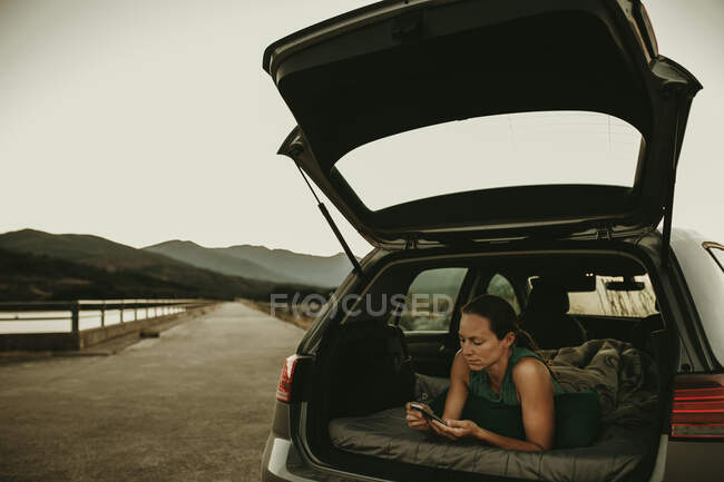 Femme utilisant un téléphone intelligent tout en étant allongé dans le coffre de la voiture contre le ciel clair — Photo de stock