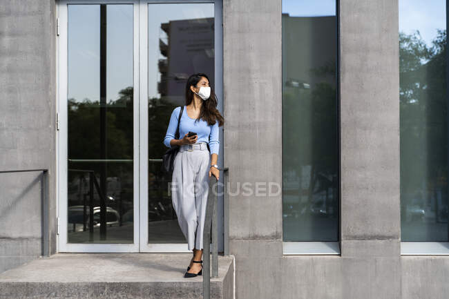 Mujer joven con mascarilla protectora de pie en la entrada del edificio durante COVID-19 - foto de stock
