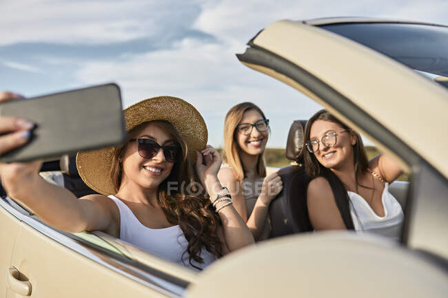Amigos tomando selfie en el teléfono inteligente mientras disfruta de viaje por carretera sentado en un coche de lujo - foto de stock