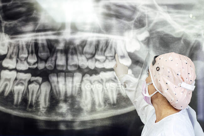 Dentiste en tenue de travail protectrice pointant vers la radiographie dentaire alors qu'il se tenait à la clinique — Photo de stock