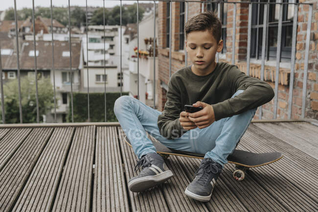 Ragazzo utilizzando smart phone mentre seduto su skateboard contro ringhiera — Foto stock