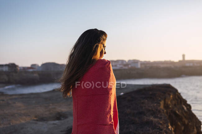 Jeune femme en châle couvert regardant la vue tout en se tenant à la plage — Photo de stock