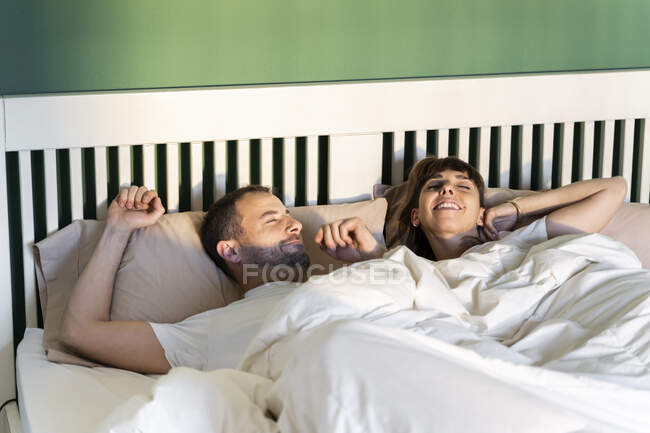 Hombre y mujer despertando lentamente mientras yacen en la cama en casa - foto de stock