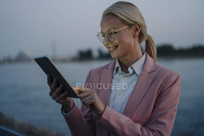 Lächelnde schöne blonde Geschäftsfrau mit digitalem Tablet gegen den Fluss in der Abenddämmerung — Stockfoto