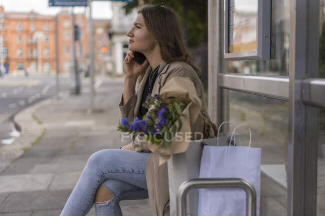 Young woman talking on smart phone with shopping bags while sitting in city — Stock Photo