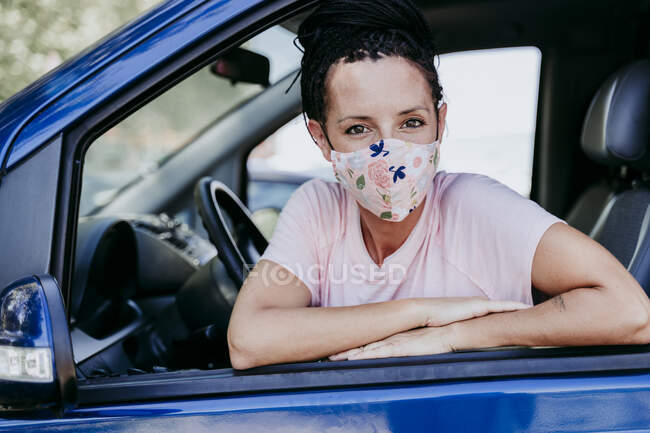 Mujer con máscara protectora floral sentada en el coche mientras se apoya en la ventana - foto de stock