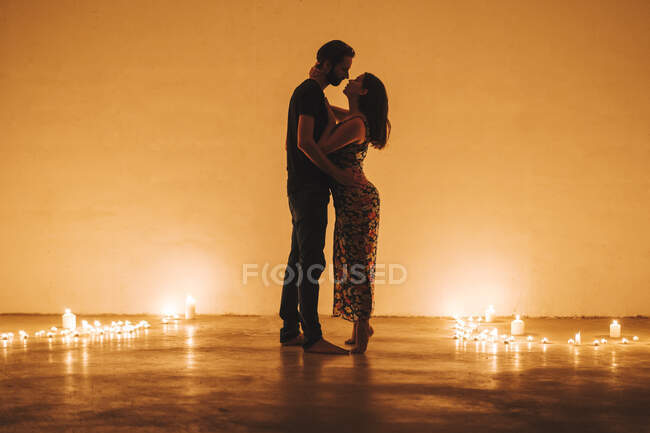 Casal romântico beijando e abraçando enquanto estava em pé em meio a velas acesas contra a parede em casa — Fotografia de Stock