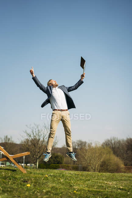 Emocionado hombre de negocios sosteniendo portátil mientras salta sobre la hierba en el parque contra el cielo despejado durante el soleado día de verano - foto de stock